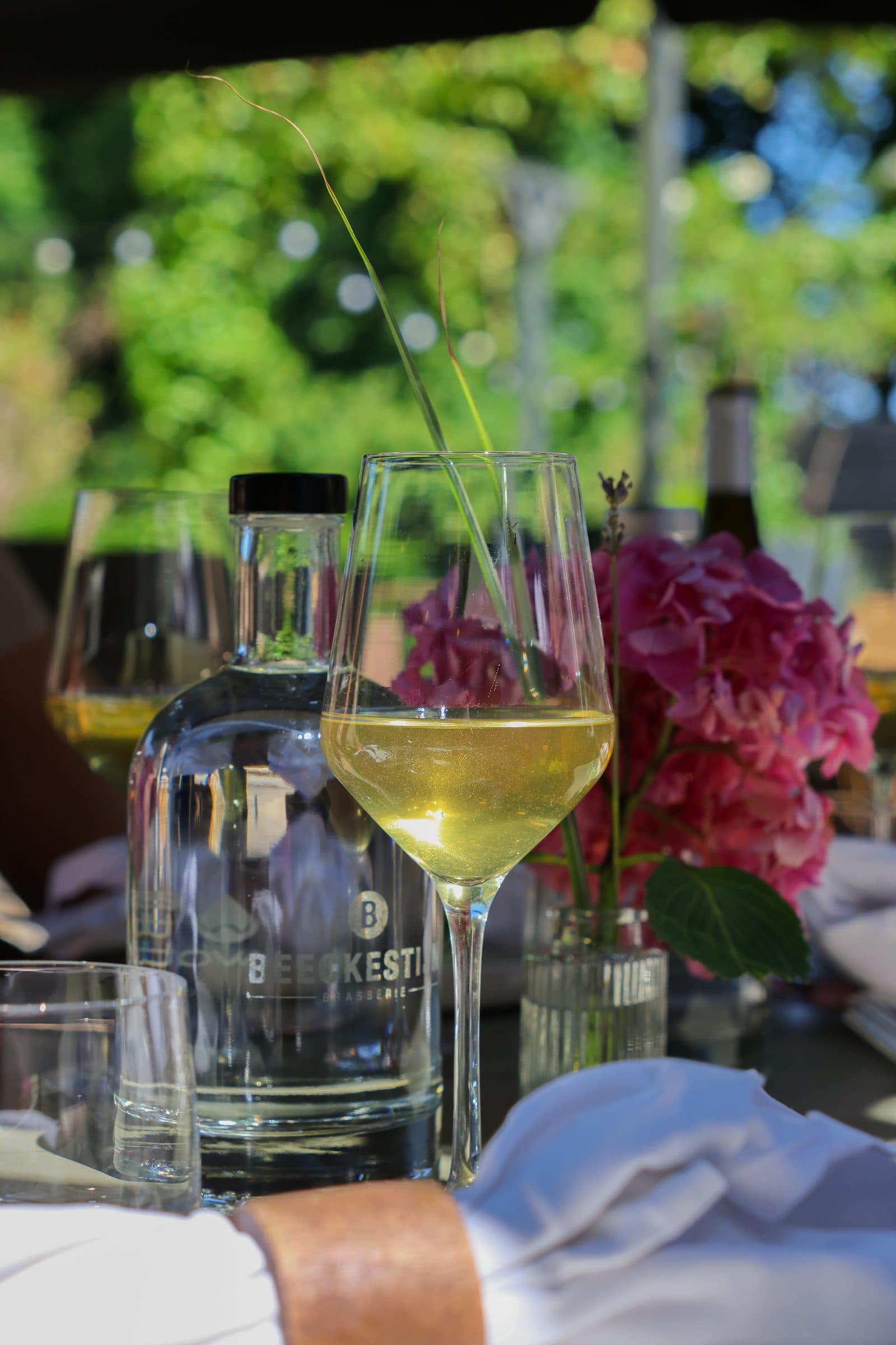 Glas witte wijn en een karaf met het logo van Beeckestijn op een gedekte tafel, met hortensia's en groen op de achtergrond.