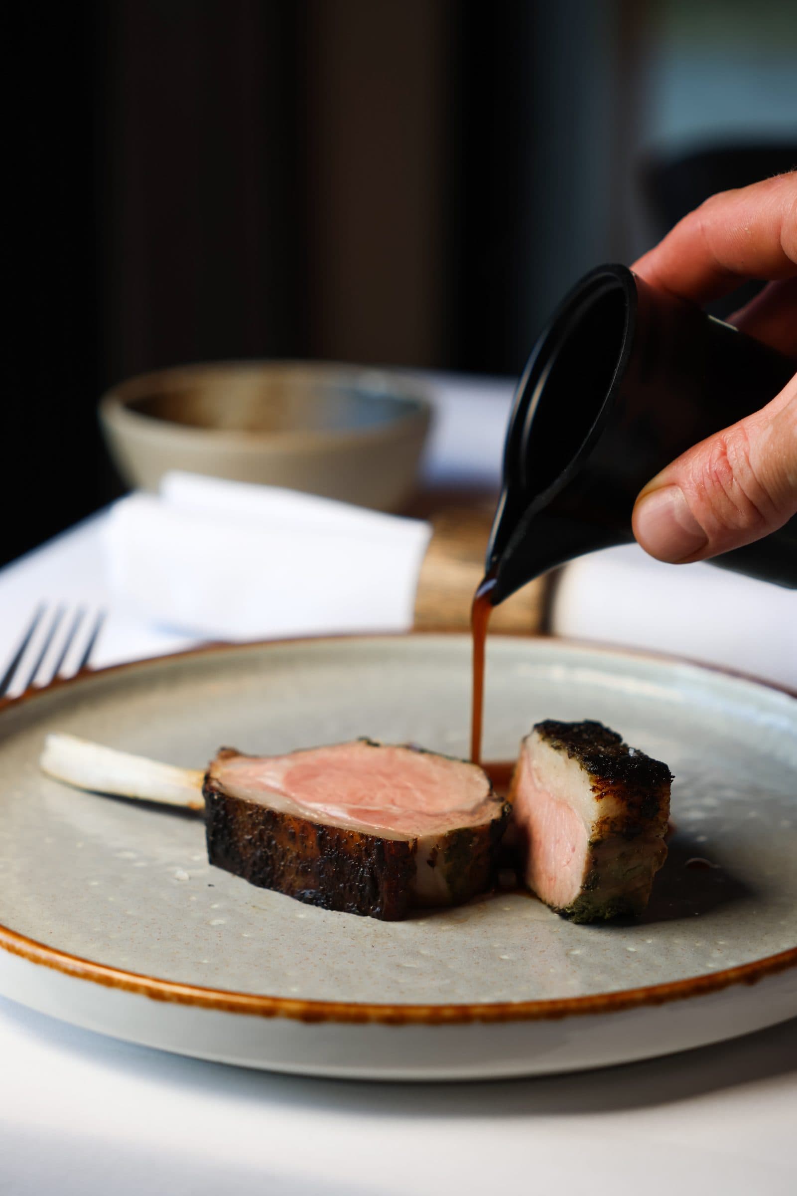 Kok schenkt een donkere jus over een rosé gebakken lamskotelet aan tafel.