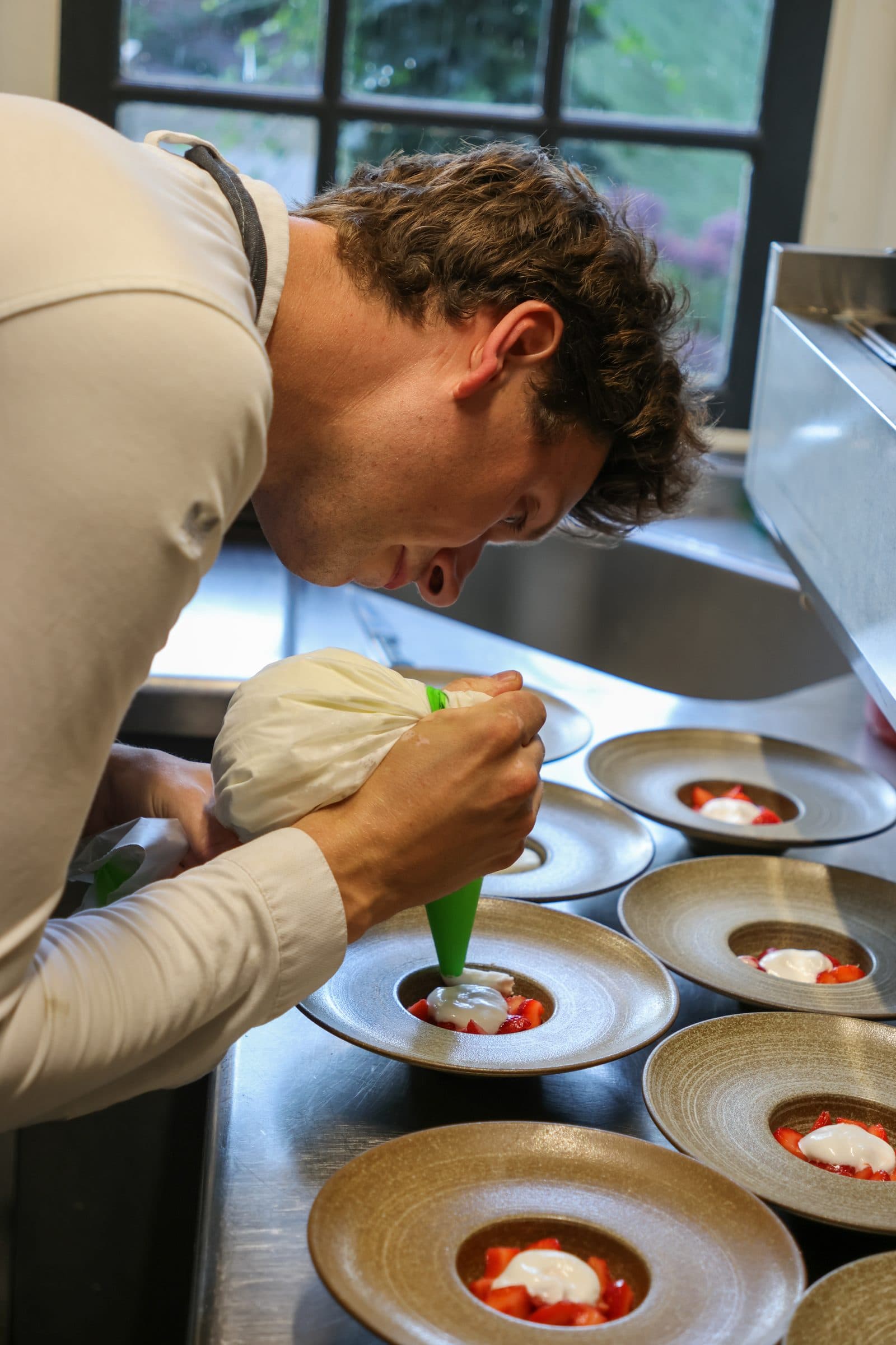 Kok spuit een crème op een dessert met verse aardbeien, in een rij borden op de keukenwerkbank.
